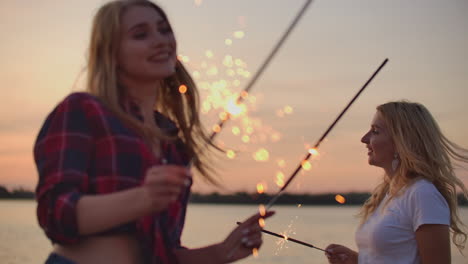 Two-girls-are-dancing-with-big-bengal-lights-on-the-river-coast.-This-is-enjoyable-summer-evening-on-the-open-air-party-at-pink-sunset.